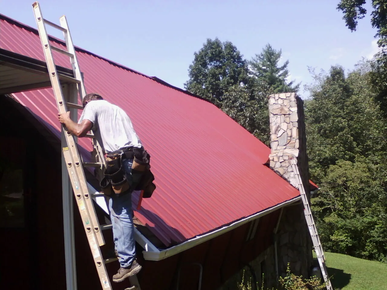 Roofer on a ladder beside a red metal roof and stone chimney — Soffit & Fascia Repair by Roofing Brothers in Tobias